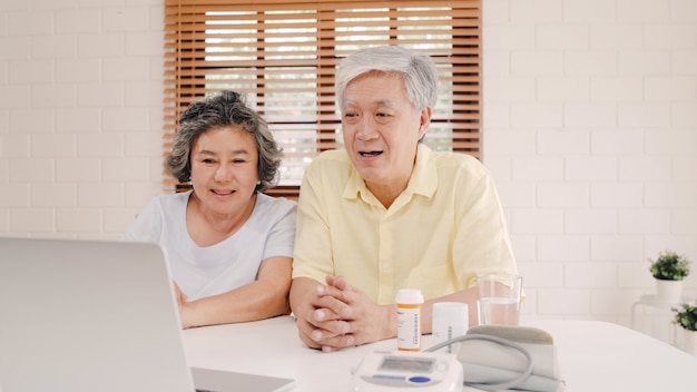 Coppie anziane asiatiche facendo uso della conferenza del computer portatile con medico circa informazioni della medicina in salone, coppia facendo uso del tempo insieme mentre trovandosi sul sofà a casa.