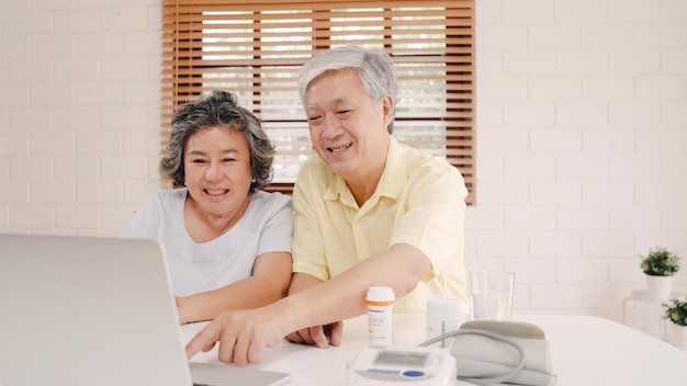 Coppie anziane asiatiche facendo uso della conferenza del computer portatile con medico circa informazioni della medicina in salone, coppia facendo uso del tempo insieme mentre trovandosi sul sofà a casa.