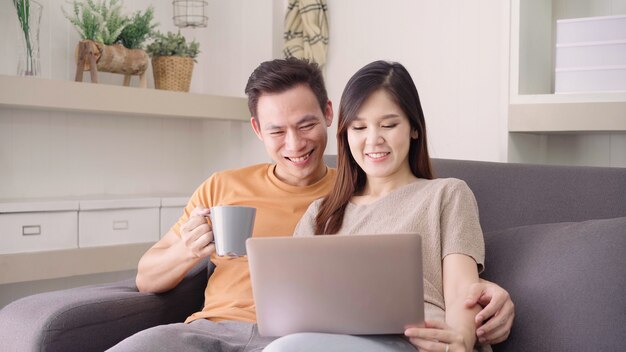 Coppie asiatiche che per mezzo del computer portatile e bevendo tazza di caffè calda in salone a casa, le coppie dolci godono di