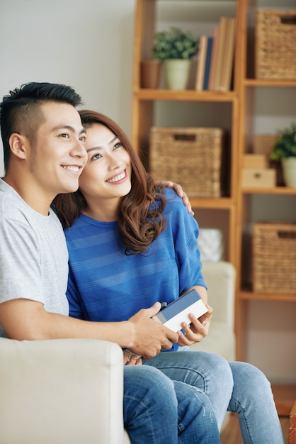 Coppie asiatiche felici che si siedono insieme sullo strato a casa, abbracciando e distogliendo lo sguardo