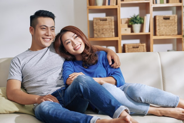 Coppie asiatiche felici che si siedono insieme sullo strato a casa, distogliendo lo sguardo e sorridendo