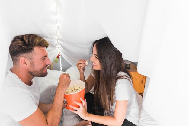 Coppie che si siedono sul letto coperto di tenda bianca che mangia popcorn