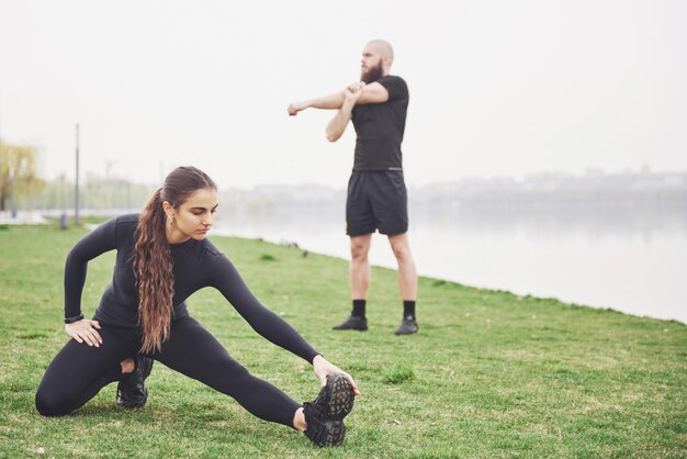 Coppie di forma fisica che allungano all'aperto nel parco vicino all'acqua. Giovane uomo e donna barbuti che si esercitano insieme nella mattina