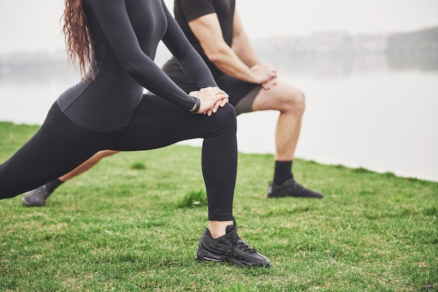 Coppie di forma fisica che allungano all'aperto nel parco vicino all'acqua. Giovane uomo e donna barbuti che si esercitano insieme nella mattina