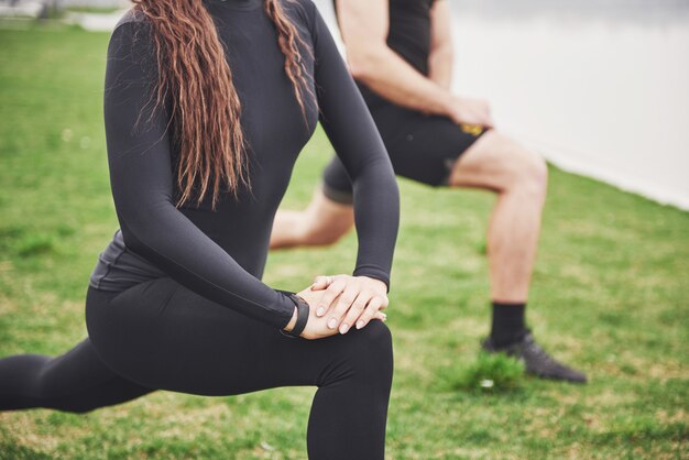 Coppie di forma fisica che allungano all'aperto nel parco vicino all'acqua. Giovane uomo e donna barbuti che si esercitano insieme nella mattina