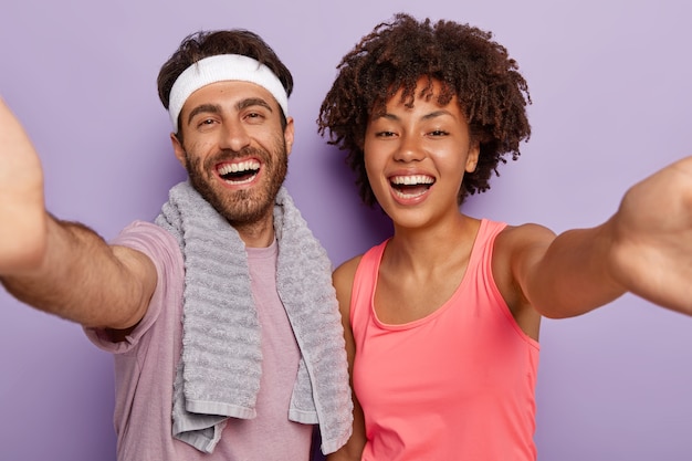 Coppie diverse positive stanno da vicino e prendono selfie, sorridono volentieri, fanno sport insieme, l'uomo sorridente indossa la fascia, l'asciugamano sul collo, sembra sano ed energico