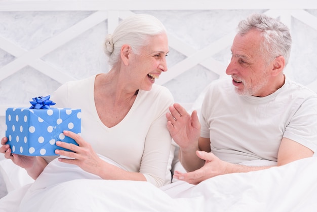 Coppie felici senior che si siedono sul letto che tiene il contenitore di regalo blu