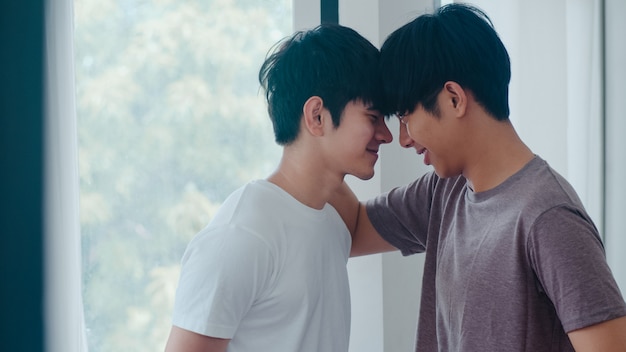 Coppie gay asiatiche che stanno e che abbracciano vicino alla finestra a casa. I giovani uomini asiatici LGBTQ + che baciano felici si rilassano insieme, trascorrono insieme momenti romantici nel salotto di casa moderna al mattino.
