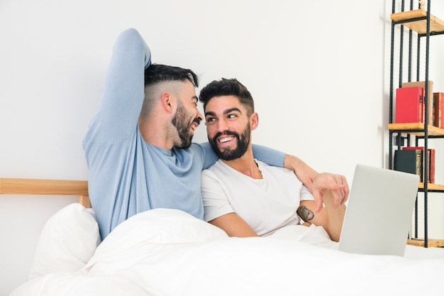 Coppie gay belle felici che si siedono insieme sul letto con il computer portatile