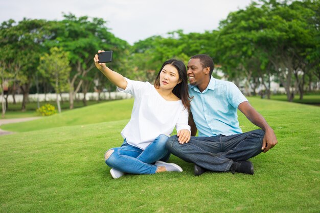 Coppie multietniche sorridenti che si siedono sul prato inglese e che prendono selfie con lo smartphone in parco.