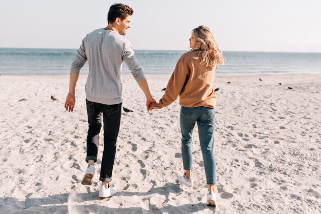 Coppie positive che corrono al mare con il sorriso. Outdoor ritratto di bella ragazza mano nella mano con il ragazzo durante il riposo in spiaggia.