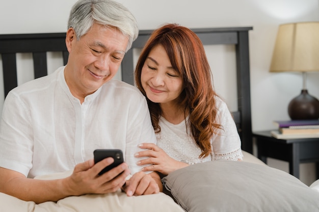 Coppie senior asiatiche facendo uso del telefono cellulare a casa. I nonni, il marito e la moglie cinesi senior asiatici felici dopo svegliano, guardando il film che si trova sul letto in concetto della camera da letto a casa di mattina.