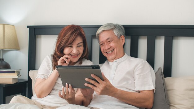 Coppie senior asiatiche facendo uso della compressa a casa. I nonni, il marito e la moglie cinesi senior asiatici felici dopo svegliano, guardando il film che si trova sul letto in concetto della camera da letto a casa di mattina.