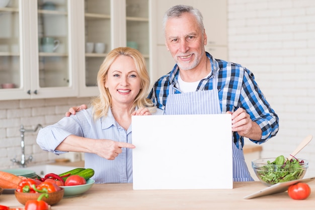 Coppie senior che mostrano cartello in bianco bianco sulla tavola di legno nella cucina