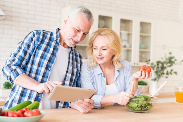 Coppie senior sorridenti che esaminano compressa digitale per la preparazione dell&#39;alimento nella cucina