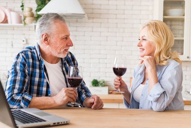 Coppie senior sorridenti che tengono vetro di vino rosso a disposizione che se lo esamina con il computer portatile sulla tavola