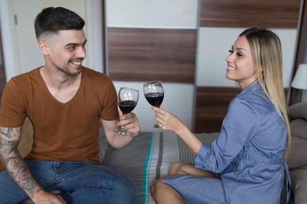 Coppie sorridenti che si siedono sul letto che tosta i bicchieri di vino