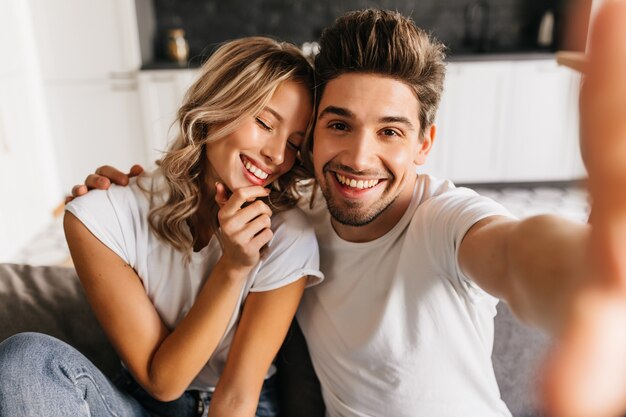 Coppie sorridenti romantiche che fanno selfie a casa seduto sul divano. L'uomo e la sua ragazza sorridono felicemente con gli occhi chiusi.