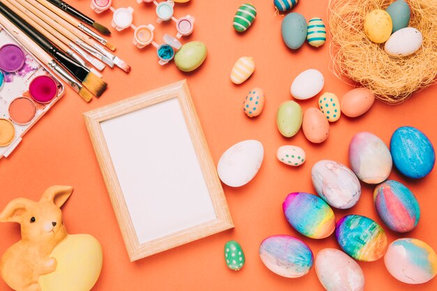 Cornice bianca vuota con uova di Pasqua colorate; pennelli; statua di acquerello e coniglio su uno sfondo arancione