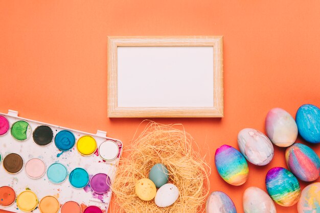 Cornice in legno bianco vuoto con scatola di pittura ad acquerello e uova di Pasqua su uno sfondo arancione