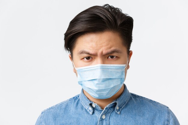 Coronavirus, distanza sociale e concetto di stile di vita. Primo piano dell'uomo asiatico arrabbiato e sconvolto in maschera medica accigliato deluso, guardando la persona che non usa misure protettive durante il covid-19.
