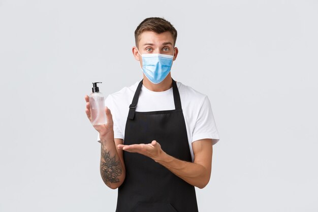 Coronavirus, distanza sociale in caffè e ristoranti, affari durante il concetto di pandemia. La maschera medica, il barista o il cameriere allegro venditore spiega l'importanza del disinfettante per le mani per la disinfezione