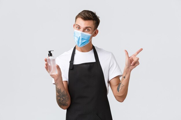 Coronavirus, distanza sociale nei caffè, concetto di pandemia aziendale. Amichevole barista tatuato in maschera medica che punta l'angolo in alto a destra per prevenire misure di virus, consiglio di usare un disinfettante per le mani