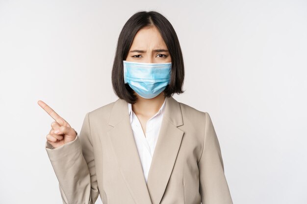 Coronavirus e concetto di lavoro Donna d'affari asiatica frustrata in maschera medica che punta il dito a sinistra e si lamenta in piedi su sfondo bianco