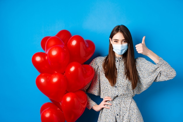 Coronavirus e concetto di pandemia. Bella donna in maschera medica e vestito in piedi vicino a palloncini di San Valentino e mostrando pollice in su, in piedi su sfondo blu
