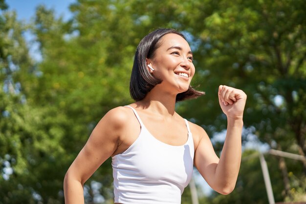 Corridore asiatico sorridente della ragazza di forma fisica in cuffie senza fili che funzionano nel parco jogging allenamento all'aperto