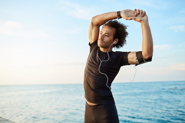 Corridore dell'uomo con l'acconciatura folta che si estende prima dell'allenamento attivo. Atleta maschio che indossa gli auricolari in abbigliamento sportivo nero facendo esercizi.