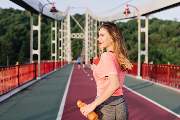 Corridore femminile beato che gode del buongiorno. Ritratto all'aperto della ragazza bianca sveglia che funziona allo stadio.