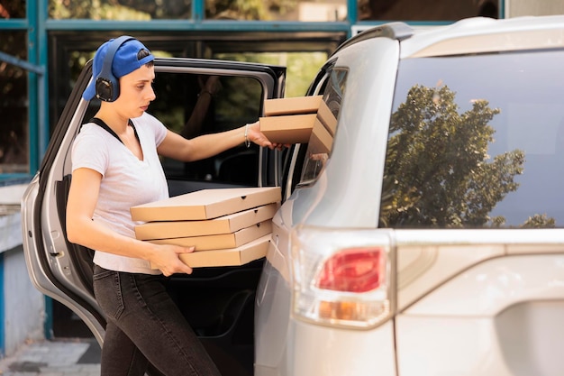 Corriere che consegna la pizza all'ufficio dalla vista laterale dell'auto, donna che tiene il mucchio di scatole. Impiegato del servizio di consegna del pranzo in cuffia che trasporta fastfood, in piedi vicino all'edificio dell'azienda all'aperto