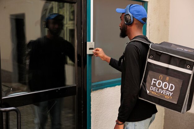 Corriere del servizio di consegna del pranzo di lavoro che suona il campanello dell'edificio per uffici, in attesa del cliente all'aperto. Giovane uomo afroamericano con zaino che consegna pasti al ristorante da asporto