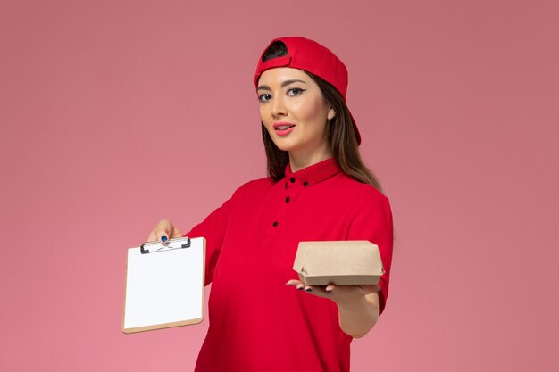 Corriere femmina giovane vista frontale in mantello rosso uniforme con piccolo pacchetto di cibo per la consegna e blocco note con la penna sulle mani sul muro rosa chiaro