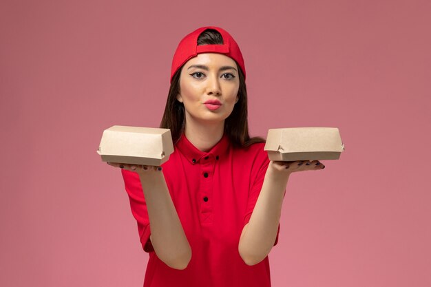 Corriere femmina giovane vista frontale in uniforme rossa e mantello con piccoli pacchi di cibo di consegna sulle sue mani sul muro rosa