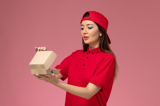 Corriere femmina giovane vista frontale in uniforme rossa e mantello con piccolo pacchetto di cibo di consegna sulle sue mani sulla parete rosa chiaro