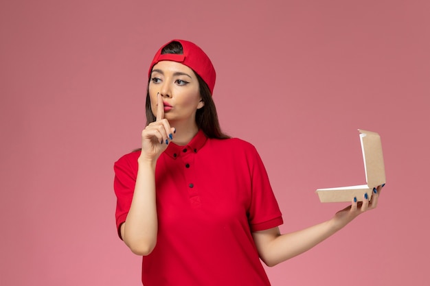 Corriere femmina giovane vista frontale in uniforme rossa e mantello con piccolo pacchetto di cibo di consegna sulle sue mani sulla parete rosa chiaro