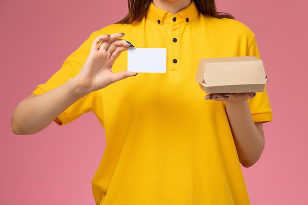 Corriere femmina vista frontale ravvicinata in uniforme gialla e mantello che tiene piccolo pacchetto di cibo per la consegna e carta sul muro rosa chiaro, lavoro di società di consegna uniforme di servizio