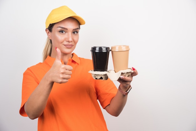 Corriere femminile biondo che tiene due tazze di caffè e che fa i pollici aumenta il segno sulla parete bianca.