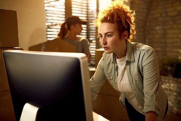 Corriere femminile che utilizza il computer e legge i dati per la consegna del pacco mentre lavora in ufficio Il suo collega è in background