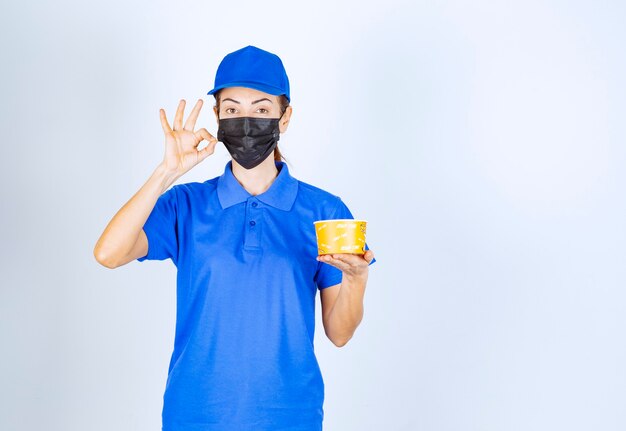 Corriere femminile del ristorante in uniforme blu e maschera facciale che consegna un cibo da asporto e garantisce il buon gusto.