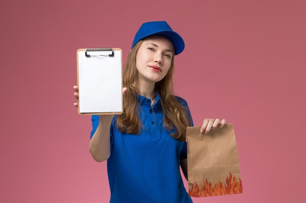 Corriere femminile di vista frontale in blocchetto per appunti della tenuta dell'uniforme blu insieme al pacchetto dell'alimento sull'azienda dell'uniforme di servizio dell'operaio di lavoro dello scrittorio rosa chiaro