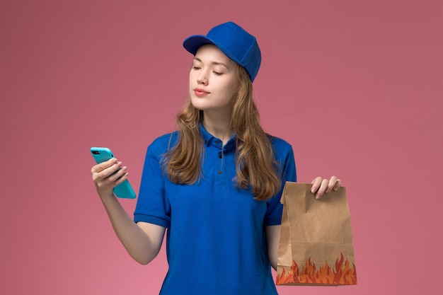 Corriere femminile di vista frontale in blu che tiene un pacchetto di cibo e telefono sul lavoro aziendale uniforme di servizio scrivania luce rosa