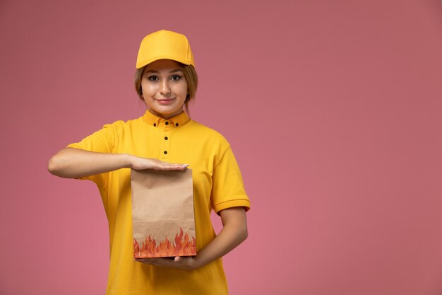 Corriere femminile di vista frontale in capo giallo uniforme giallo che tiene il pacchetto di consegna del cibo sul lavoro di lavoro di consegna uniforme sfondo rosa