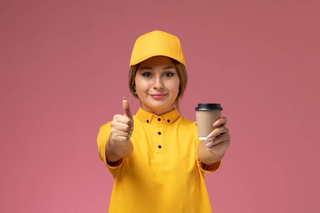 Corriere femminile di vista frontale in capo giallo uniforme giallo che tiene la tazza di caffè di plastica sulla femmina della foto di colore del lavoro di consegna uniforme del fondo rosa