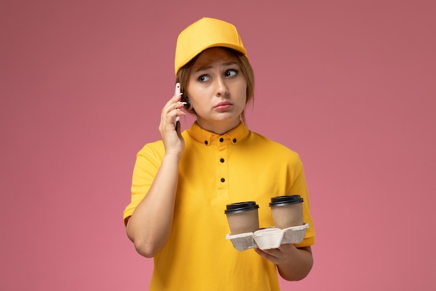 Corriere femminile di vista frontale in capo giallo uniforme giallo che tiene le tazze di caffè di plastica che parlano sul telefono sul lavoro di colore del lavoro di consegna dell'uniforme rosa della scrivania