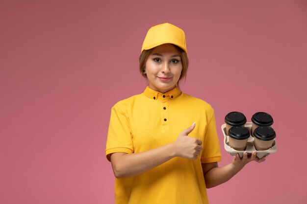 Corriere femminile di vista frontale in capo giallo uniforme giallo che tiene le tazze di caffè di plastica che sorridono sul lavoro di colore del lavoro di consegna uniforme del fondo rosa