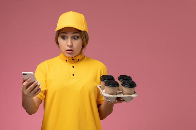 Corriere femminile di vista frontale in capo giallo uniforme giallo che tiene tazze di caffè di plastica e telefono bianco su lavoro di colore del lavoro di consegna uniforme del fondo rosa