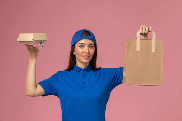 Corriere femminile di vista frontale in capo uniforme blu che tiene diversi pacchetti di consegna sulla parete rosa, lavoratore di consegna dipendente di servizio